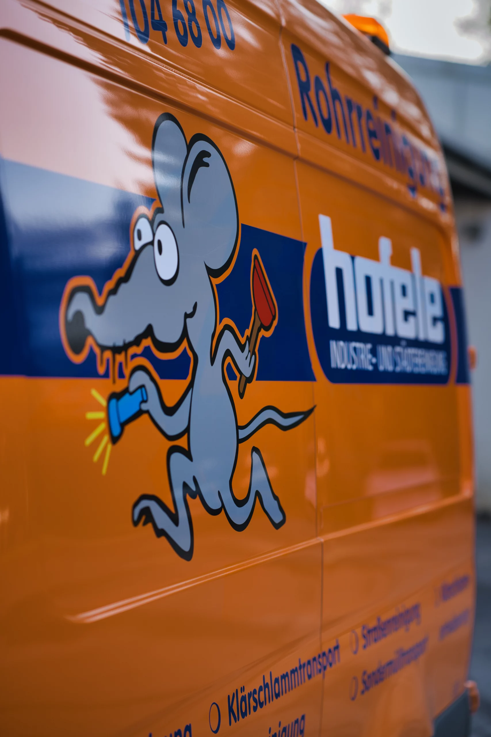 Nahaufnahme des Hofele-Firmenlogos mit Comic-Maus-Maskottchen Detailansicht der orangefarbenen Fahrzeugseite mit dem blauen Hofele-Logo und der Rohrreinigungs-Comic-Maus.